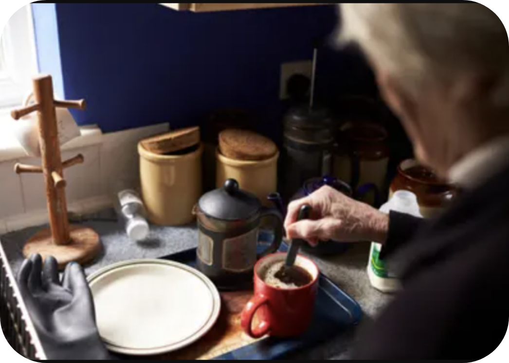 An old person standing stirring a cup of coffee with Tea ware around her