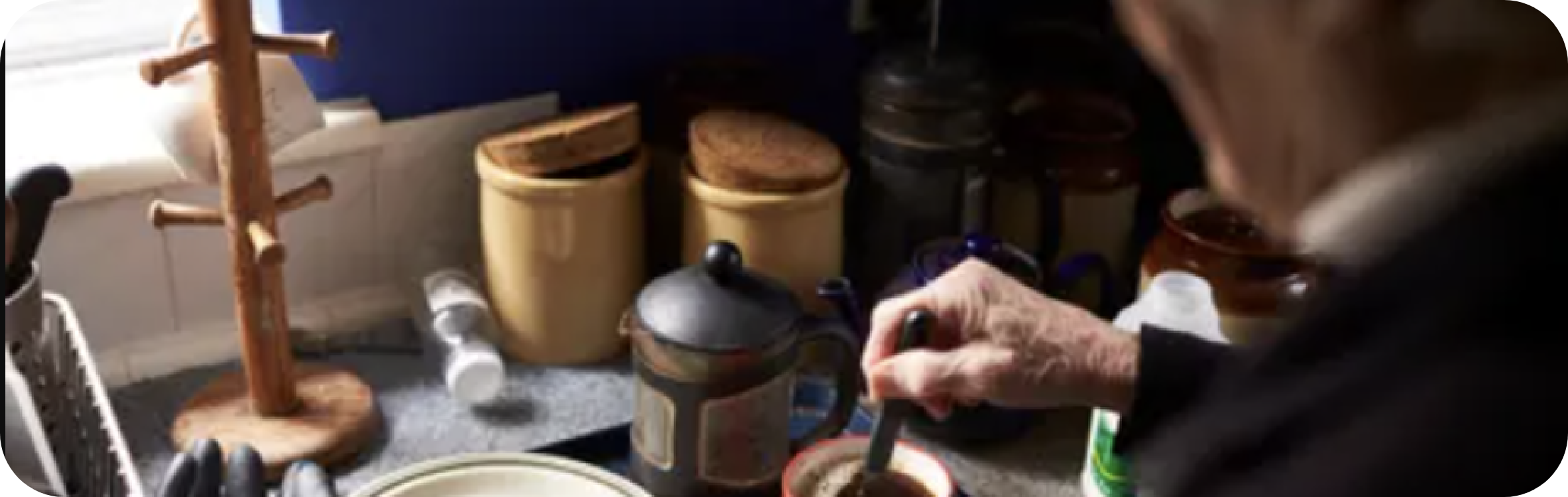 An old person standing stirring a cup of coffee with Tea ware around her